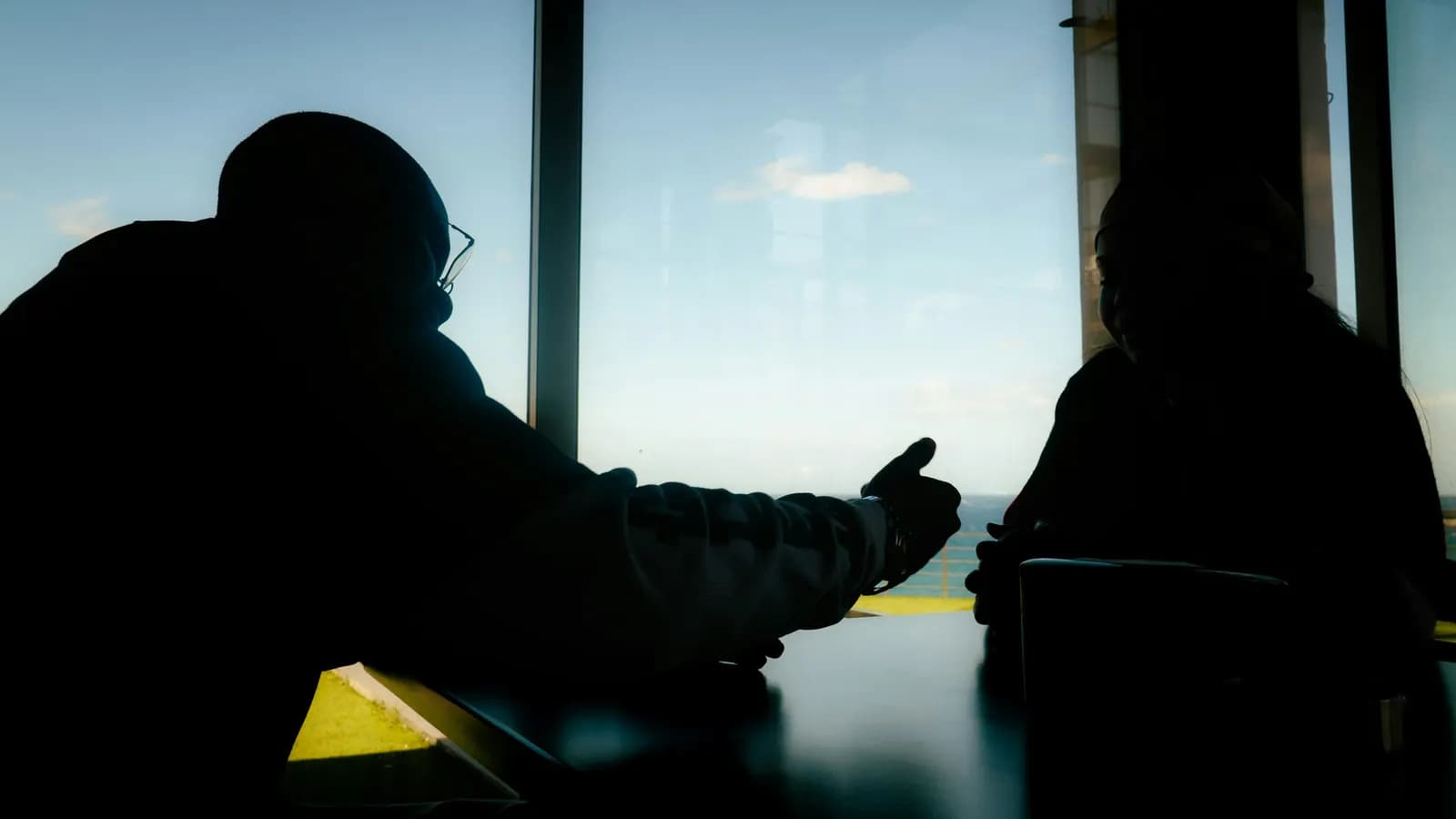 Two people in silhouette talking across a table in front of a large, bright window