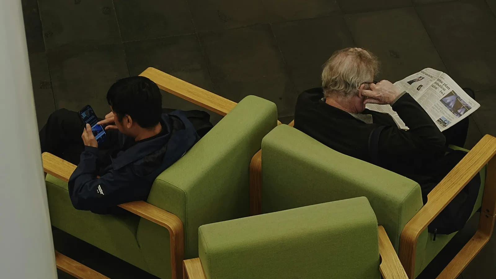 Two people in green armchairs, seen from above, one reading a newspaper and one using a phone