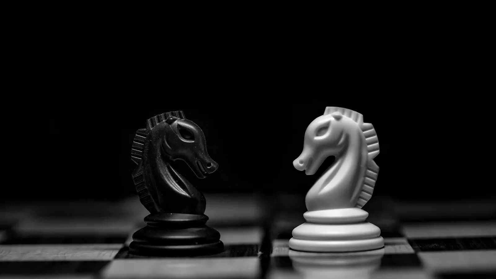 A black and a white chess knight facing each other on a dark board