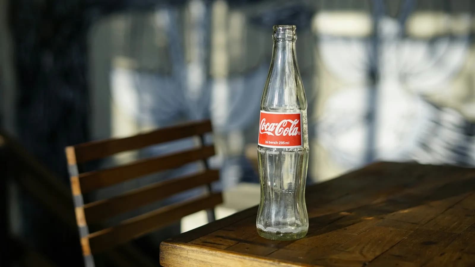 A glass Coca-Cola contour bottle on a wooden café table beside a slatted chair