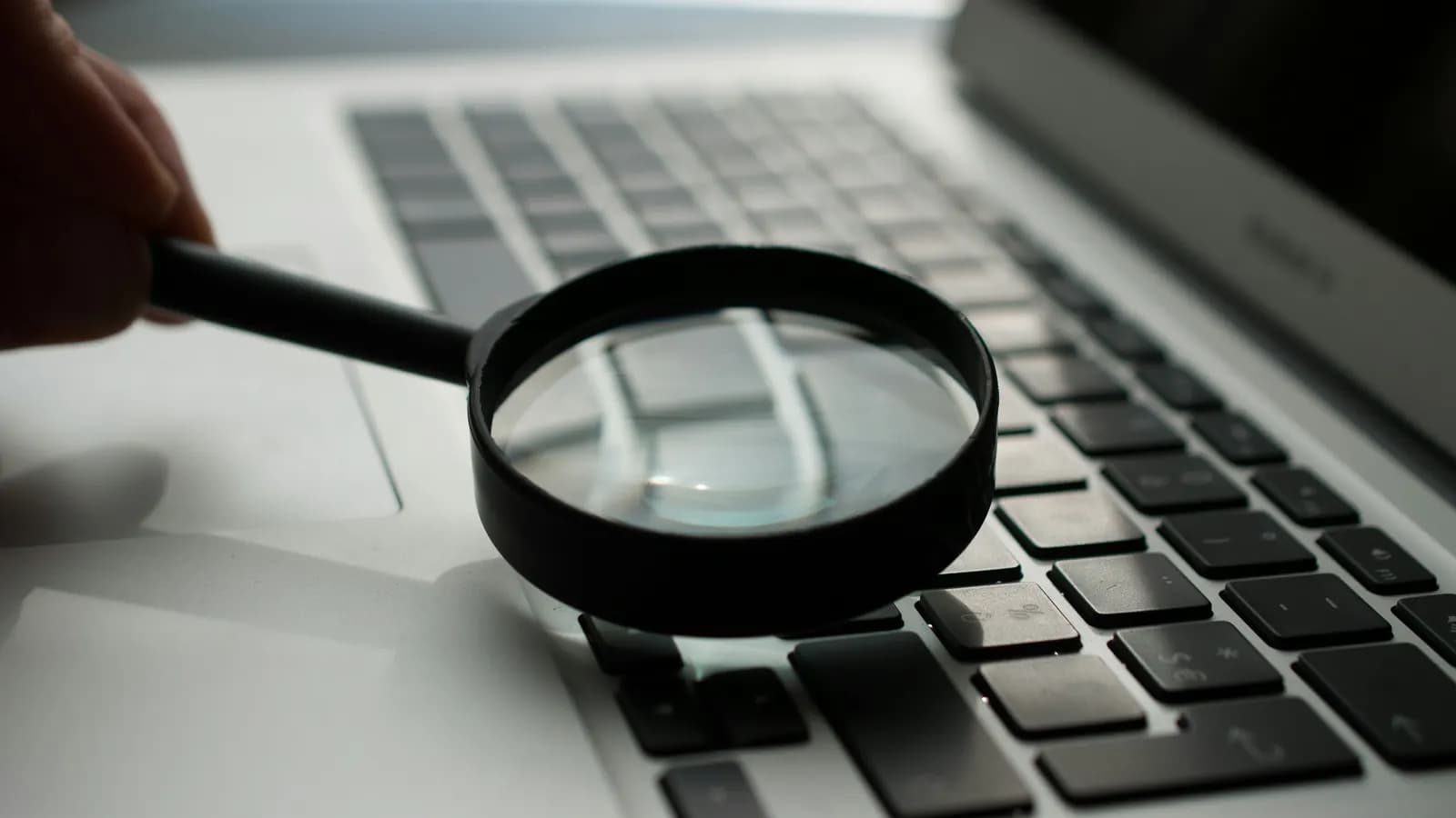 A hand holding a magnifying glass over a laptop keyboard