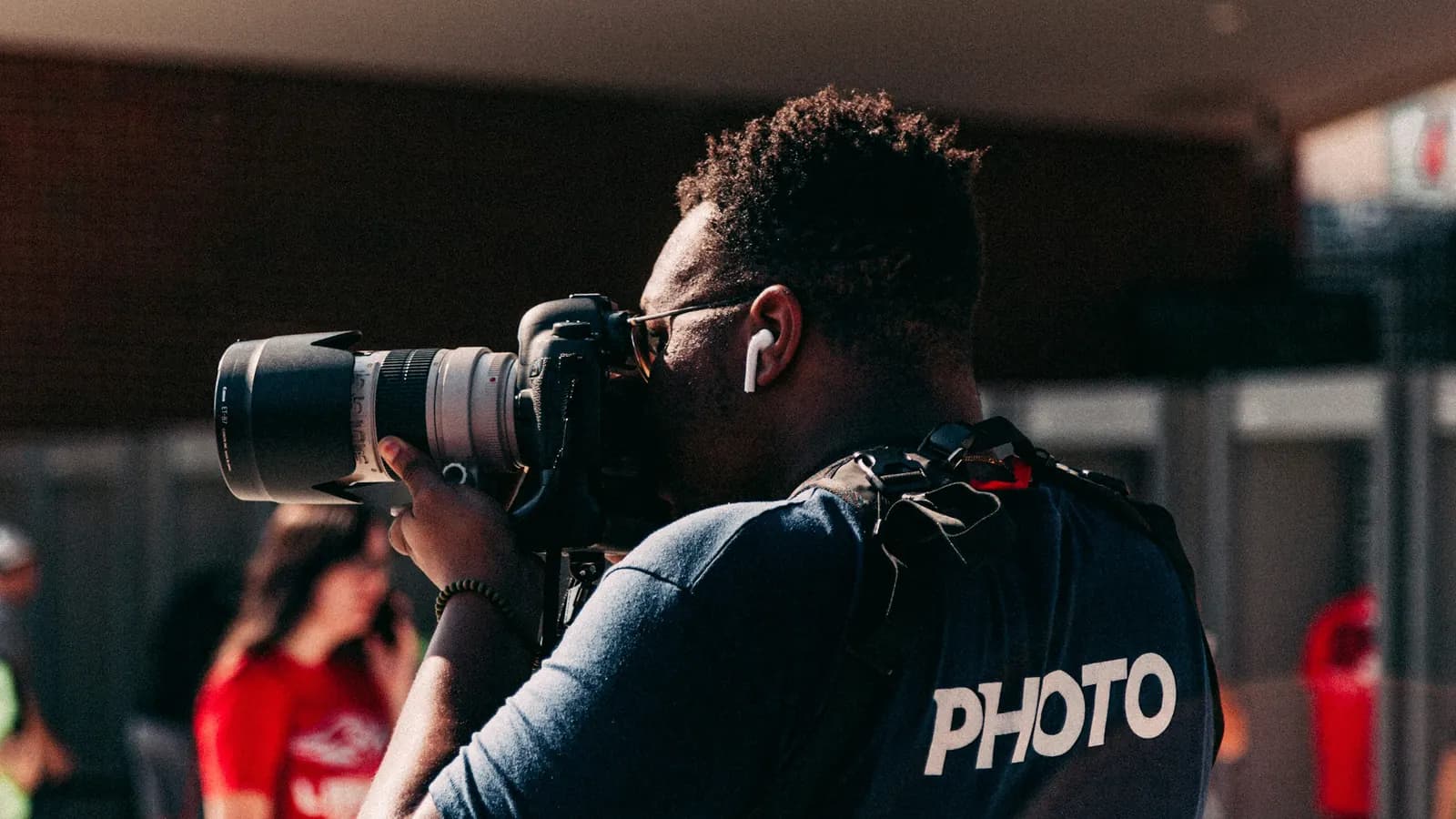 A press photographer in a PHOTO shirt, shooting with a long telephoto lens